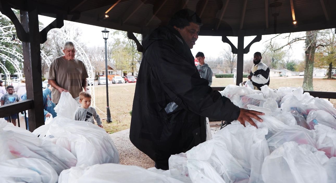 People distributing food packages under a pavilion in a community park.