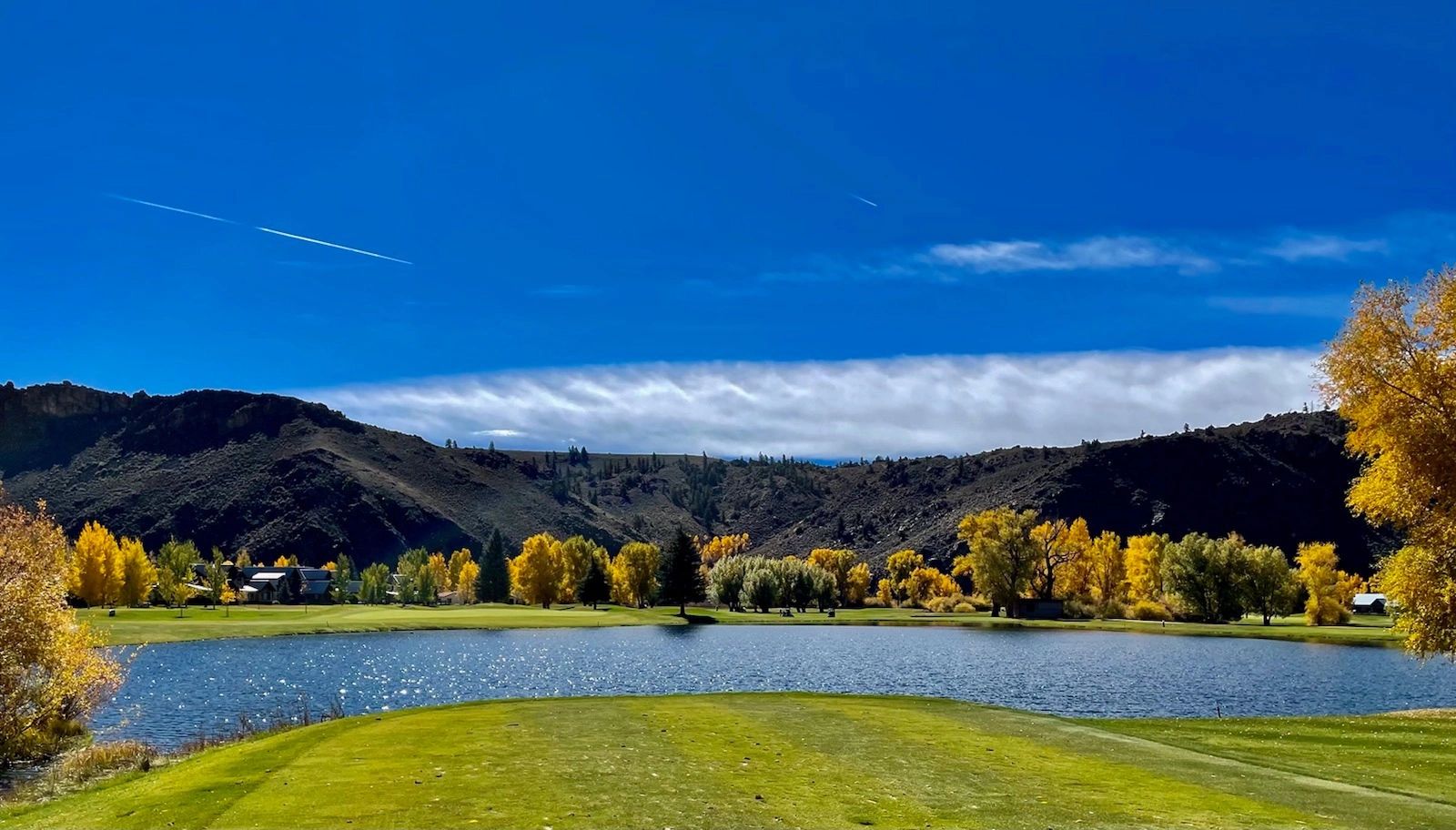 Scenic golf course by a lake with autumn trees and hills under a clear blue sky.