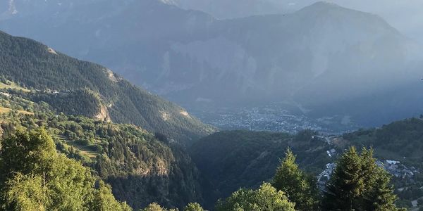 Sunlit mountain valley with lush green trees and distant town under a partly cloudy sky.