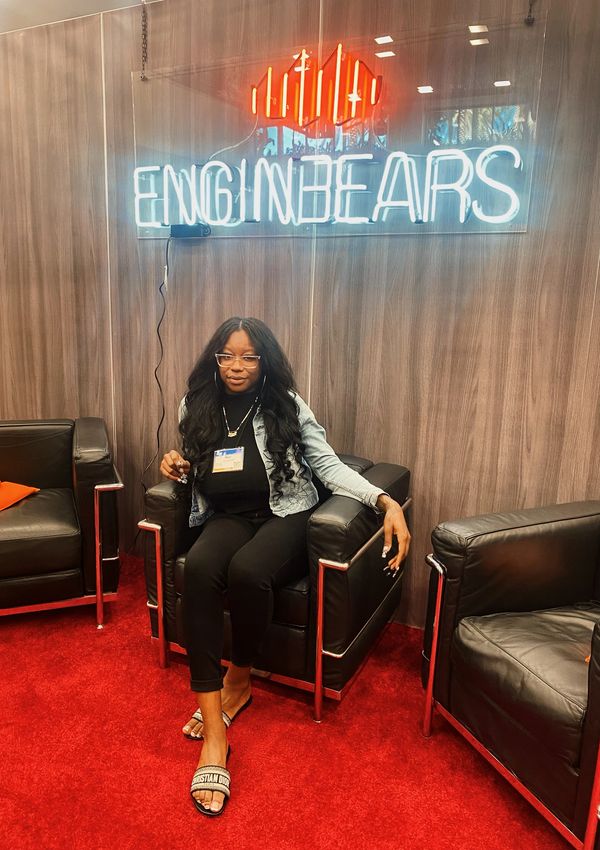 A woman sitting on a black leather chair under a neon sign that says 'ENGINBEARS'.
