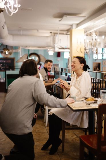 A woman sitting at a café table smiles at a man kneeling beside her, with a waiter in the background.