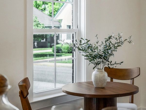 Minimalist dining nook with wooden table, chairs, and a vase with greenery by the window.