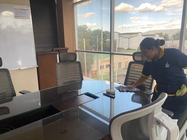 A woman cleaning a glass conference table in a bright office room.