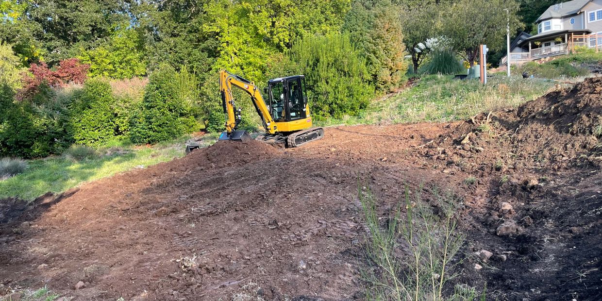 Yellow excavator working on a dirt clearing in a backyard near trees and houses.