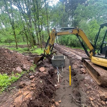 Excavator digging a trench in a forested area with concrete structures and pipes.