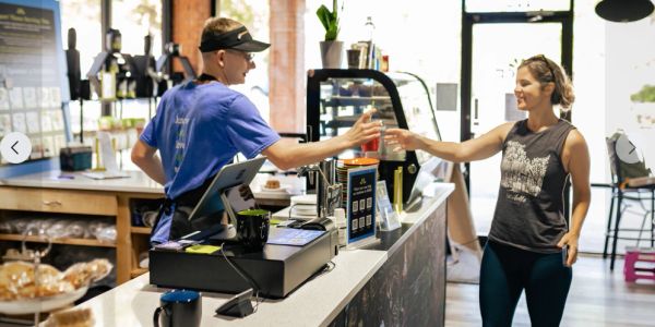 Barista handing a drink to a smiling female customer at a coffee shop counter.