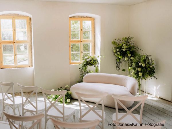 Bright room with white chairs, a curved sofa, and floral arrangements near windows.