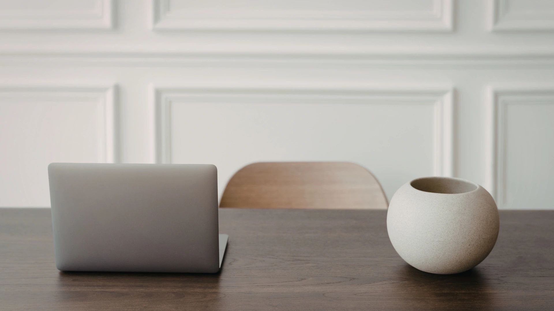 Minimalist workspace with a closed laptop and round ceramic pot on a wooden table.