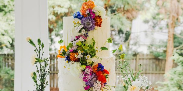 Two-tier white cake adorned with cascading colorful flowers on a blue glass stand.