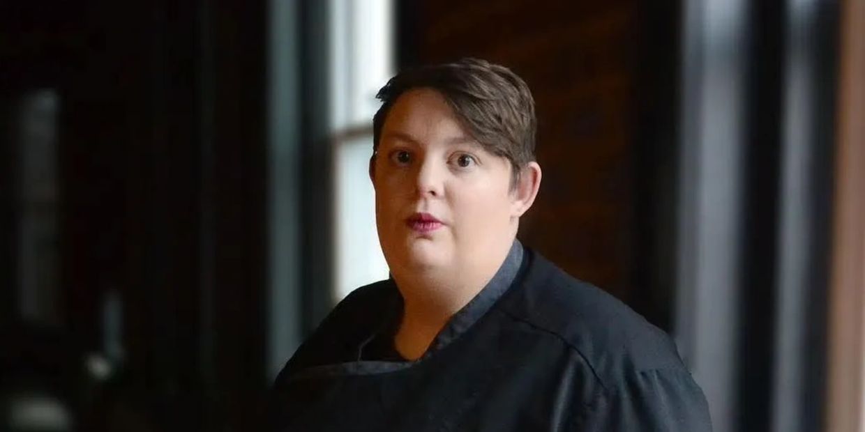 Executive chef in a black uniform standing indoors near a window.