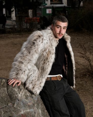 Man in a fur coat leaning on a rock outdoors.