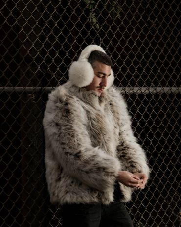 Man wearing fur coat and earmuffs against chain-link fence.