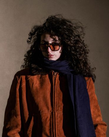 Person with curly hair wearing brown jacket and dark scarf.