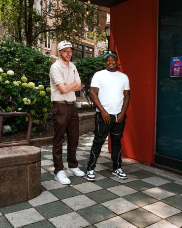 Two men standing outside by a building and plants.