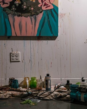 Paint jars and tools scattered below a canvas with drips on the wall.