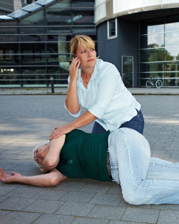 A woman calls for help while comforting an unconscious man lying on the ground.