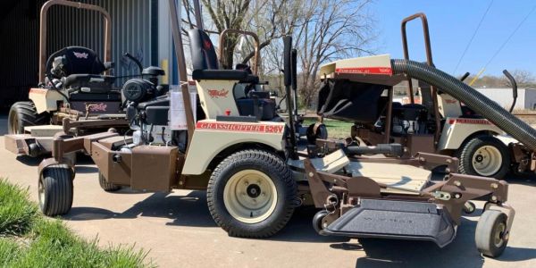 Two vintage Grasshopper lawn mowers parked on a driveway in front of a building.
