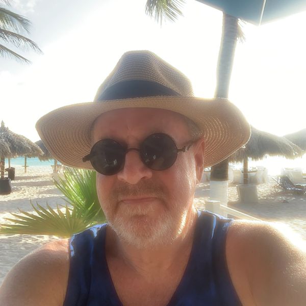 Man with round sunglasses and straw hat at a sunny beach resort.