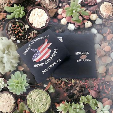 Two black T-shirts with patriotic and bar themes on a succulent-covered surface.
