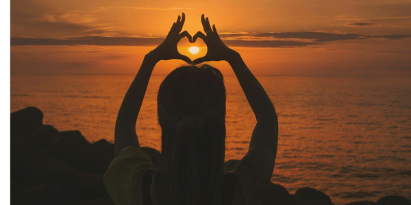 a teenager creating a heart around the sun with her hands and overlooking the ocean