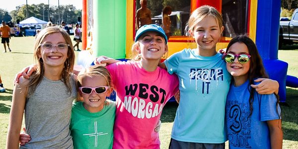 Five smiling kids posing outdoors at a sunny event.