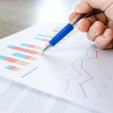 Hand holding a blue pen pointing at colorful bar and line charts on paper.