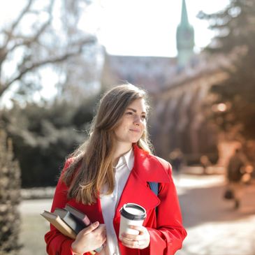 Young woman in a red coat holding books and a coffee cup outdoors.