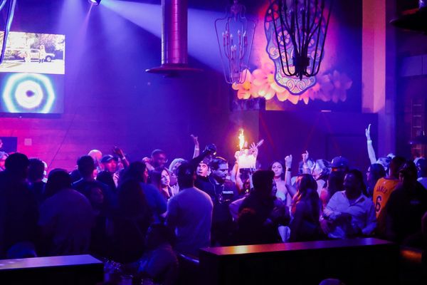 Crowded nightclub with vibrant purple lighting and people celebrating with drinks and sparklers.