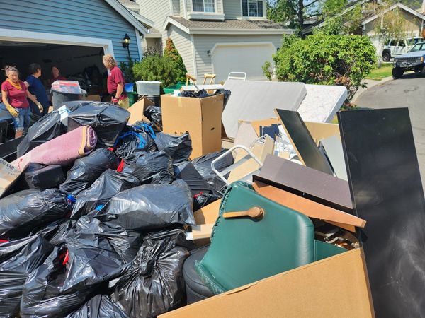 People cleaning out a garage with large piles of trash and discarded furniture outside.
