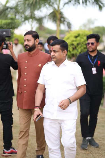 Two men walking outdoors, one in white and one in brown traditional attire.