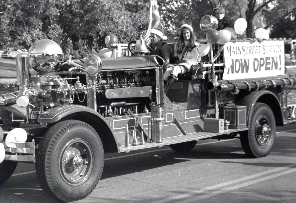 Vintage fire truck parade float with people and a 'Main Street Station Now Open!' sign.