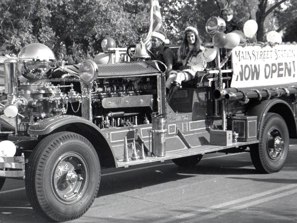 Vintage fire truck parade float with people and a 'Main Street Station Now Open!' sign.
