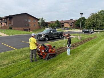 Two men operate and observe a small tracked machine on a grassy slope.