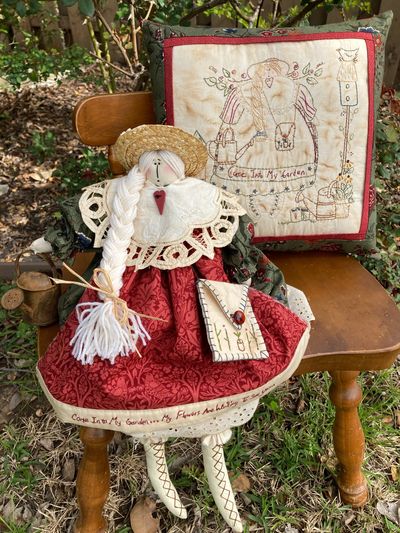 A doll on a chair next to an embroidered pillow