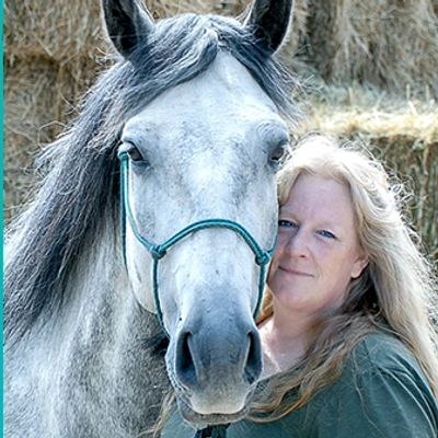 Woman posing with a gray horse in front of hay.
