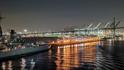 Night view of a lit-up port with cranes and a docked ship reflecting on water.