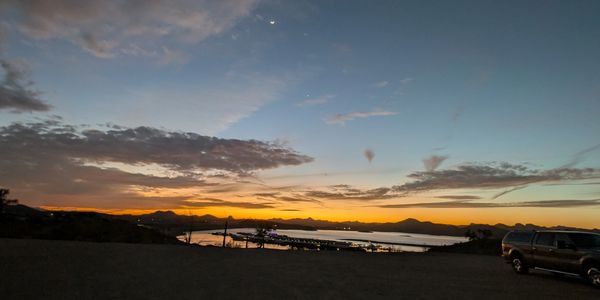 Sunset over a calm bay with mountains and a parked truck.