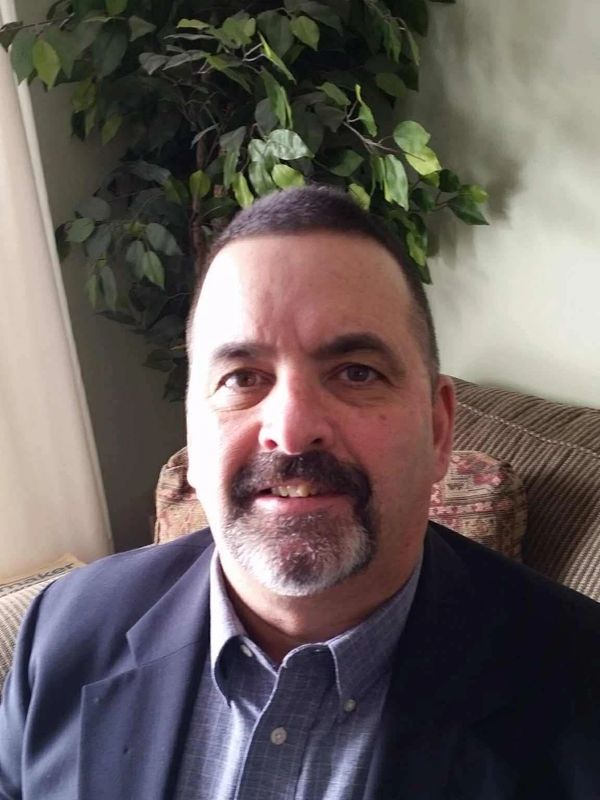 Middle-aged man smiling indoors with a plant in the background.