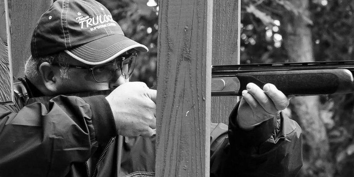 Man aiming a shotgun behind a wooden post.