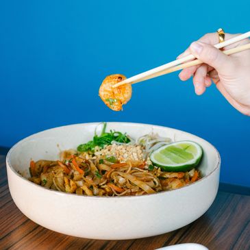 Hand holding chopsticks lifting shrimp from a bowl of noodles with lime.