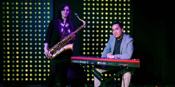 A woman holding a saxophone stands to the left of a man sitting behind a Nord keyboard. 