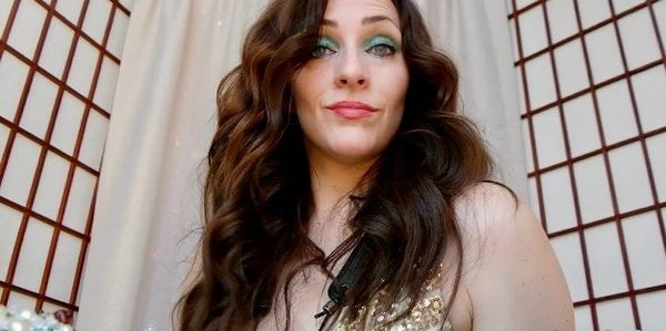 A woman with long curly brown hair and green eye shadow stares directly into the camera, shot slight