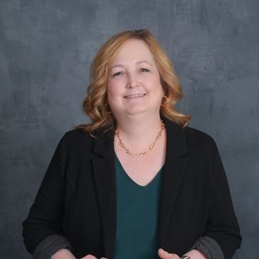 Smiling woman in a black blazer and green top against a gray backdrop.