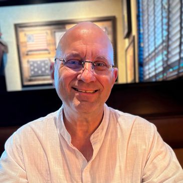 Smiling bald man with glasses wearing a white shirt in a warmly lit room.