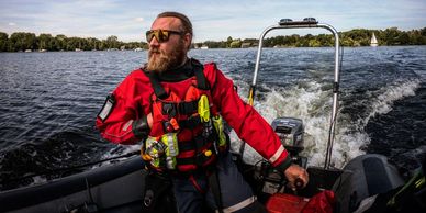 Man steering a boat on a lake wearing a red life jacket and sunglasses.