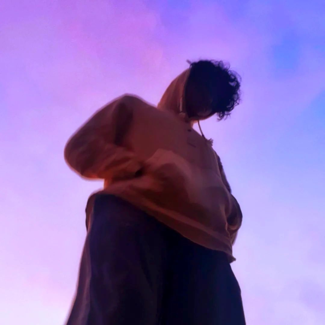 A hooded figure looking down, hands in pocket infront of a Lavender and Blue sky