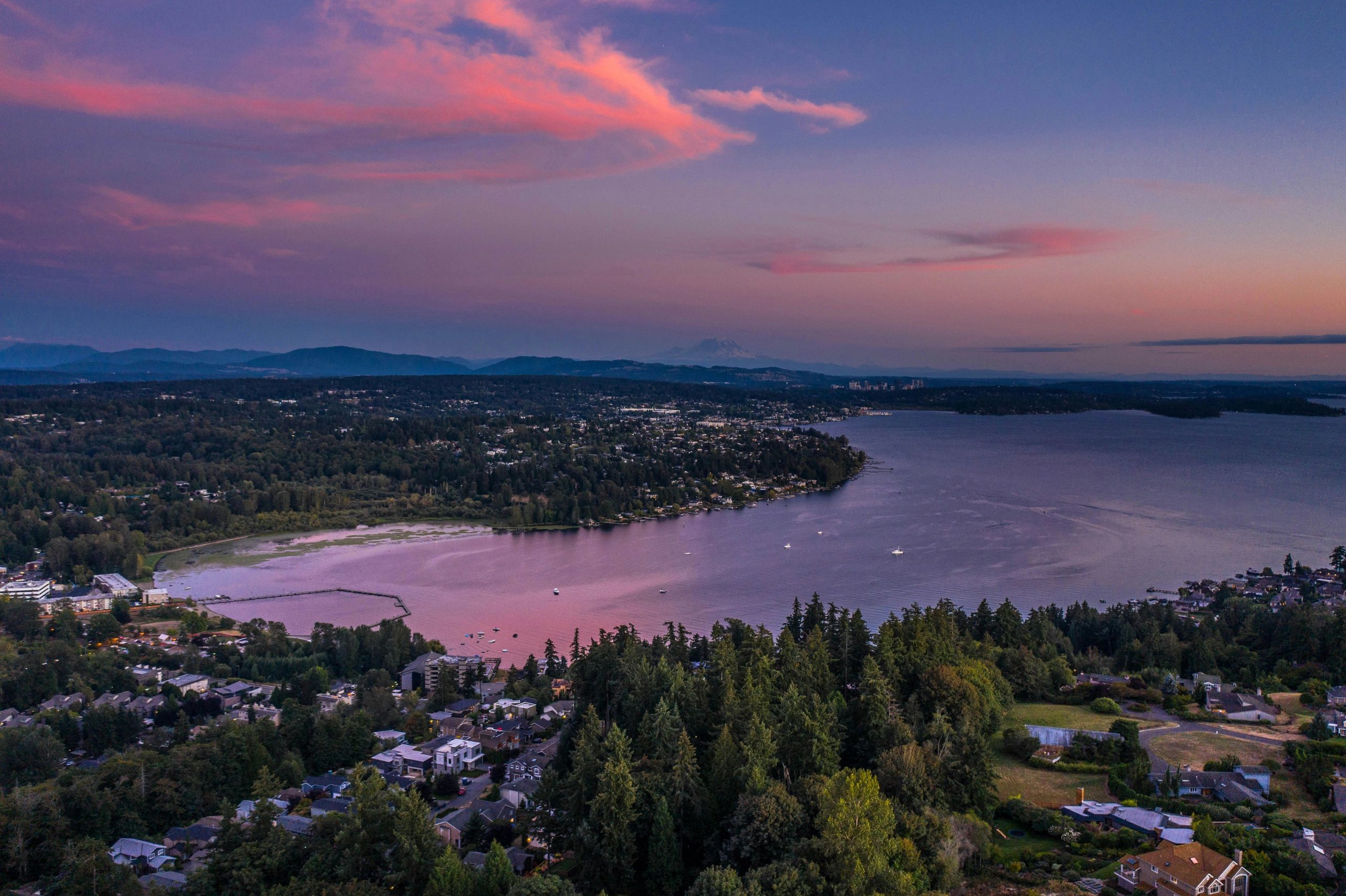 A serene lakeside town at sunset with pink clouds and surrounding forest.