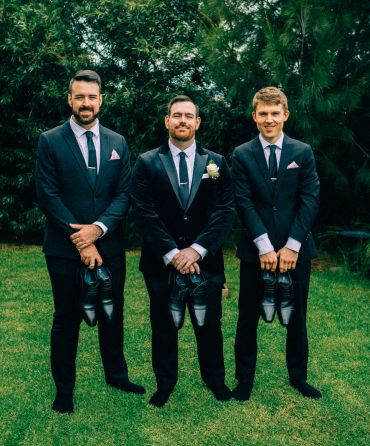Three men in suits holding shoes, posing outdoors.
