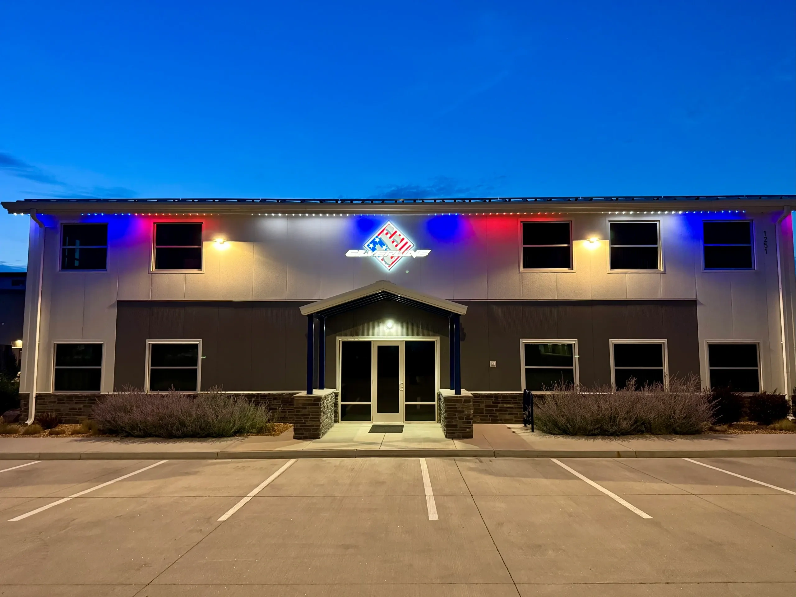 Front entrance of a building with colorful lights and a logo at dusk.