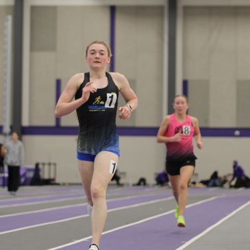 Female athletes sprinting indoors on a track during a race.
